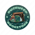 A circular sticker with the logo of Duckweed Bookstore: a greenish-blue swamp with a small frog wearing a yellow shirt laying beneath a large open book with a picture of the planet Earth on the cover. There are fairy lights strewn about the inside of the cover. The text is placed around the inside of the circular sticker and reads "Duckweed Bookstore©" with two small emblems between the words resembling lilypads.